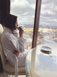 Side view of thoughtful woman drinking coffee while looking through window at home