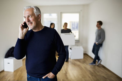 Mature man using mobile phone while family with moving boxes in background at home