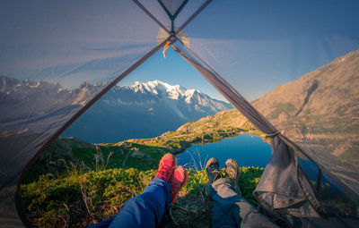 Crop pleasant hikers in colorful sneakers lying with crossed legs in small transparent tent near clear lake in mountains in sunny day in chamonix, mont-blanc