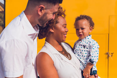 Interracial family of three smiling in front of yellow wall