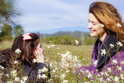 Mother with daughter sitting on grassy field amidst flowers against sky