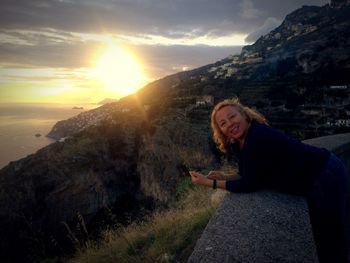 Portrait of smiling woman leaning on retaining wall at mountain during sunset
