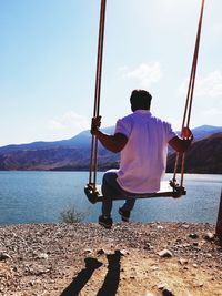 Rear view of man sitting on swing by lakeshore against sky