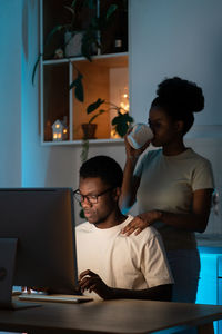 Young african woman supporting husband while he coding on computer and working remotely overtime