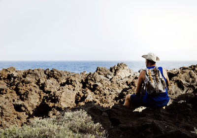 Rear view of woman sitting on rock at beach against clear sky