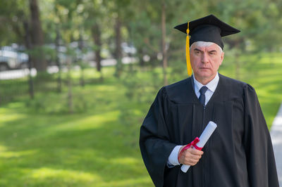 Serious old man in graduation gown holding diploma outdoors