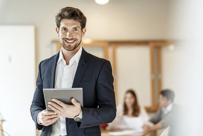 Portrait of confident businessman holding tablet with a meeting in background