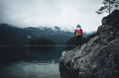 Rear view of man standing on rock at lakeshore against mountains