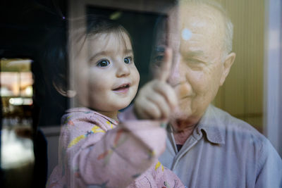 Close-up of cheerful granddaughter touching window while being carried by grandfather at home