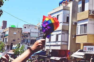 Cropped hand holding rainbow flags by buildings against sky on sunny day