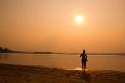 Rear view of woman standing at beach against sky during sunset