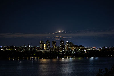 Illuminated buildings by river against sky at night