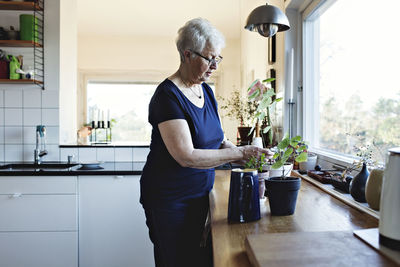 Retired senior woman standing by potted plants on kitchen counter at home