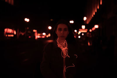 Portrait of young woman with illuminated cable standing on street in city at night