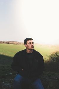 Portrait of young man sitting on field against sky