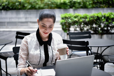 Businesswoman writing in book with laptop on table while sitting at sidewalk cafe