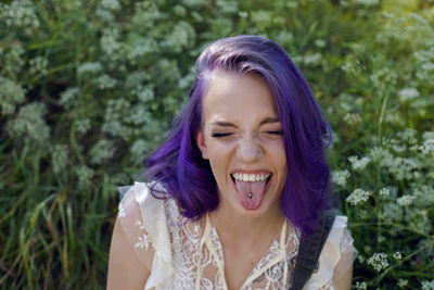 Portrait informal teenage girl with purple hair and piercing shows tongue on green field in summer