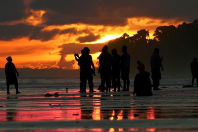 Silhouette people standing on beach against orange sky