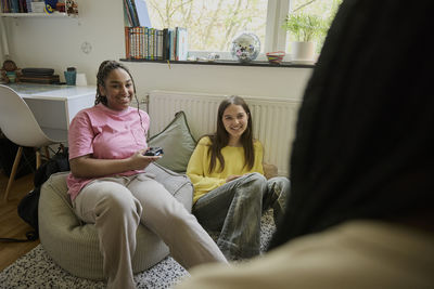 Smiling teenage girl holding game controller while talking with female friends at home
