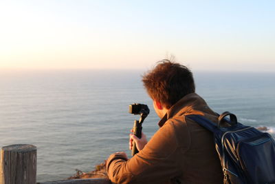 Rear view of man photographing sea against sky during sunset