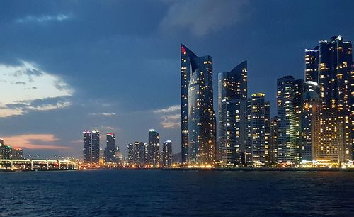 Illuminated modern buildings by bay against sky at night