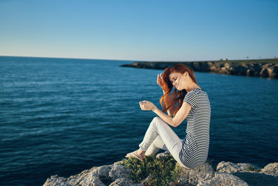 Woman sitting on rock by sea against clear sky