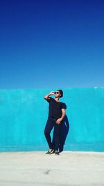 Full length of young woman wearing sunglasses posing by wall against clear blue sky during sunny day