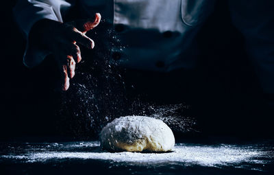 Midsection of chef sprinkling flour on dough at table in dark
