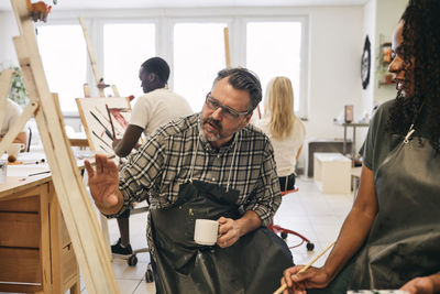 Male instructor explaining female student over artist's canvas in art class