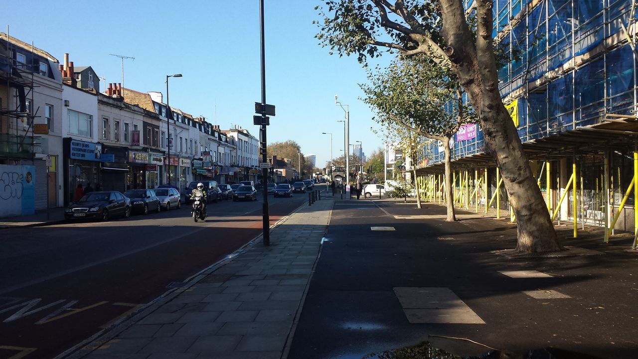 building exterior, architecture, built structure, city, street, the way forward, tree, transportation, road, street light, car, sunlight, clear sky, incidental people, sky, city street, city life, blue, road marking, shadow