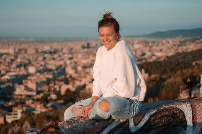 Portrait of smiling woman sitting on rock against cityscape