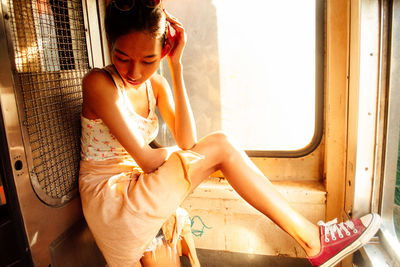 Young woman looking away while sitting on window at home