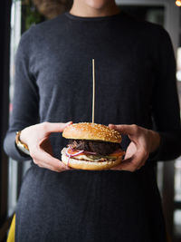 Crop anonymous person in black wear holding classic burger with cutlet and vegetables with cheese while representing fast food industry