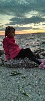 Man sitting on shore at beach against sky during sunset