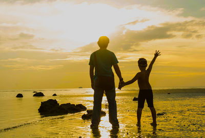 Father and son standing on beach against sky during sunset