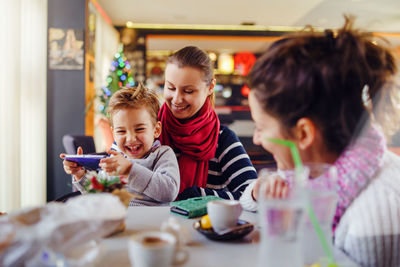 Friends looking at cheerful boy playing with video game in restaurant