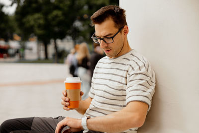 Young adult man sitting outdoors, drinking coffee and waiting for someone. man looking at his watch