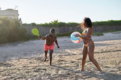 Cheerful african american male playing tennis near black girlfriend with colorful ball while spending time on sandy beach on sunny day