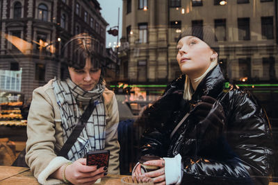 Thoughtful young woman looking through glass window while friend using mobile phone in cafe during winter