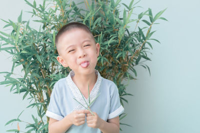 Cute boy sticking out tongue while standing against plants and wall