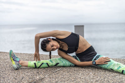 Fit female athlete sitting on embankment and doing side bend while stretching muscles and warming up during training