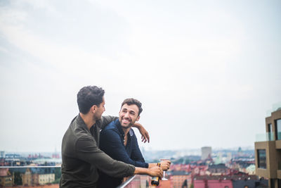 Happy man looking at male friend with drink while leaning over glass railing on rooftop against sky