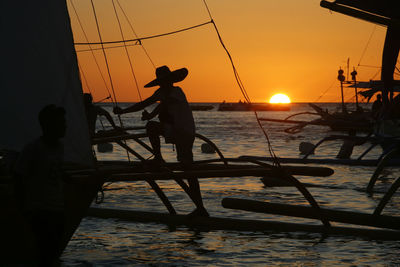 Man climbing on outrigger boat in sea against sky during sunset