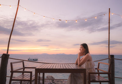 Side view of woman sitting by sea on bench during sunset