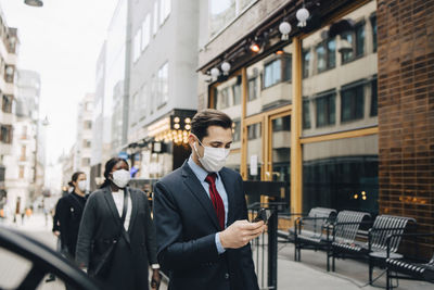 Businessman using smart phone while female colleagues social distancing in city during pandemic