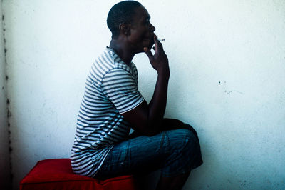 Side view of man smoking cigarette while sitting by wall