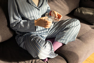 Girl is sitting on the sofa, holding a glass of water and a blister pack with pills in her hands