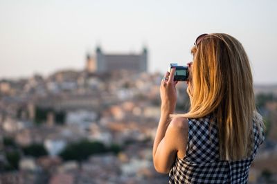 Rear view of girl photographing buildings using camera on sunny day