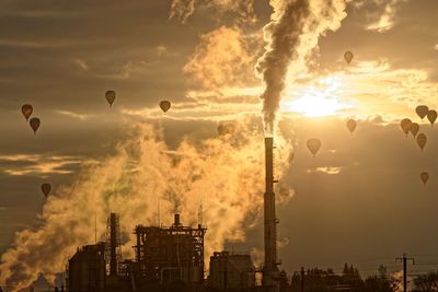 Silhouette hot air balloons flying over factory during sunset