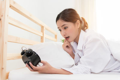 Young woman holding alarm clock while lying on bed at home